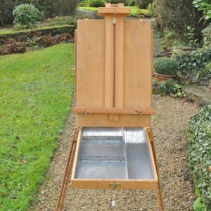 May include: A wooden artist's easel with a pull-out drawer and a metal tray. The easel is set up outdoors on a gravel surface.