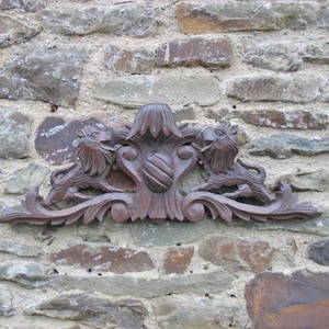 May include: A carved wooden decorative element featuring two lion-like creatures flanking a central shield design. The dark brown piece is mounted on a rough stone wall, showcasing intricate details and a classic aesthetic.