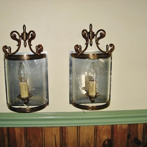 May include: Two vintage wall sconces with a brass finish. Each sconce has a clear glass enclosure with a candle-shaped light bulb and a decorative brass top. The sconces are mounted on a cream-coloured wall above a green and wood-panelled surface.