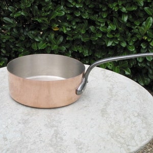 May include: A copper saucepan with a long, silver handle. The saucepan is sitting on a light gray, textured surface.