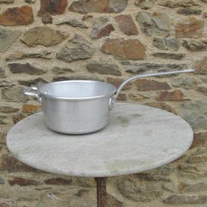 May include: A silver aluminium colander with a long handle sits on a round, light grey stone table. The colander has small holes throughout the bottom.