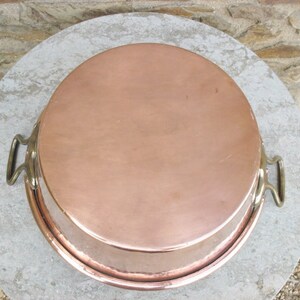 May include: A large, round copper baking pan with two brass handles. The pan is sitting on a light gray surface.