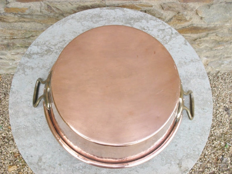 May include: A large, round copper baking pan with two brass handles. The pan is sitting on a light gray surface.