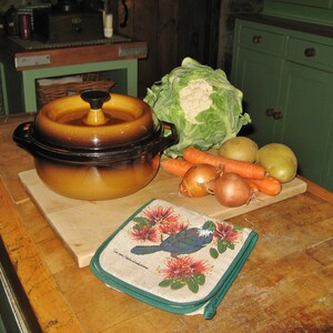 Puede incluir: Una olla de cerámica marrón con tapa se encuentra sobre una encimera de madera junto a una tabla de cortar con verduras. Las verduras incluyen una cabeza de coliflor, dos cebollas, tres zanahorias y dos patatas. También hay un agarrador de olla floral verde y blanco en la encimera.