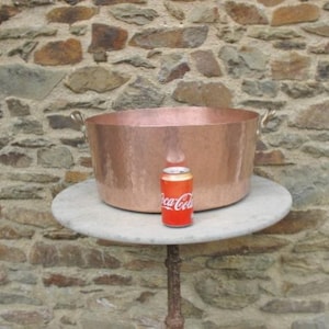 May include: A large copper pot with two handles sits on a white marble table. A can of Coca-Cola is in front of the pot for scale.