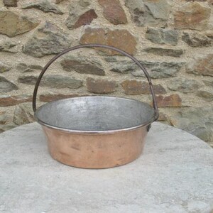 Pode incluir: Um balde de cobre e metal com uma alça de metal curva. O balde tem formato redondo e fica sobre uma superfície redonda de cor clara. O fundo é uma parede de pedra.