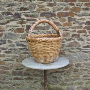 May include: A woven wicker basket with a single handle sits on a white marble table with a black metal base. The basket is light brown and has a natural, rustic look.