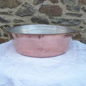 May include: A large, antique copper bowl with a wide, shallow design. The bowl has a polished copper exterior and a silver-colored interior. It features two brass handles and sits on a white cloth, with a stone wall in the background.