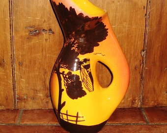 May include: A yellow and orange ceramic pitcher with a brown silhouette of a tree and a bird. The pitcher has a handle and a wide mouth.