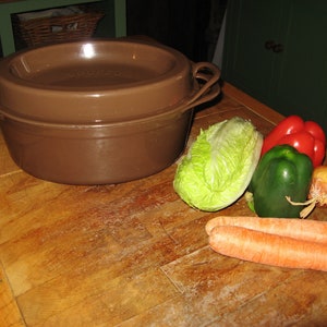 Könnte beinhalten: Ein brauner gusseiserner Topf mit Deckel, der auf einer Holzoberfläche steht. Neben dem Topf liegen Gemüse, darunter ein Salatkopf, zwei rote Paprika, ein grüner Paprika, zwei Zwiebeln und zwei Karotten.