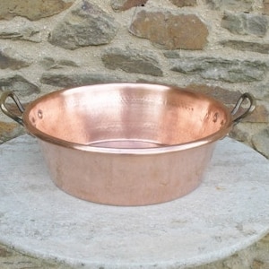 May include: A large, round copper basin with two handles. The basin is sitting on a light gray stone surface.