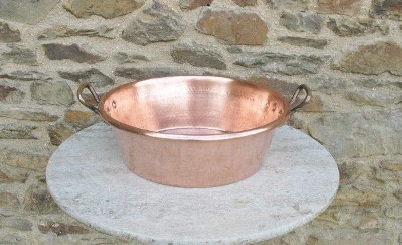 May include: A large, round copper basin with two handles. The basin is sitting on a light gray stone surface.