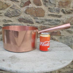 May include: A large, polished copper saucepan with a long, straight handle and a distinct pouring spout. The pan is positioned next to a standard red and white aluminum Coca-Cola soda can, which serves as a visual reference for its size.