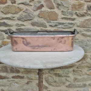 Puede incluir: Un molde para hornear rectangular de cobre con dos asas de hierro. El molde está sobre una mesa de mármol blanco.