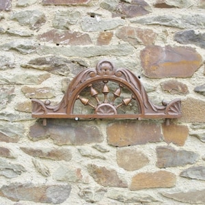 May include: A decorative wooden wall mount with a carved design featuring a wheel with spokes and a curved top. The mount is attached to a stone wall.