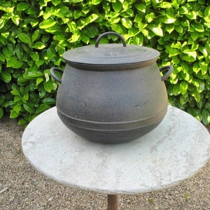 以下が含まれることがあります： 白い丸いテーブルの上に、蓋付きの大きな黒い鋳鉄製の鍋が置かれています。鍋には3つの取っ手と、わずかに丸みを帯びた底があります。