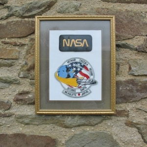 May include: A framed image of a NASA patch with the text "NASA" in gold lettering. The patch is surrounded by a smaller patch depicting the space shuttle Challenger with the names of the crew members: McNAIR, ONIZUKA, RESNIK, SCOBEE, SMITH, McAUliffe, and JARVIS.