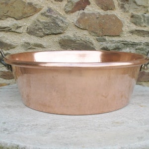 May include: A large copper bowl with two brass handles. The bowl is round and has a smooth, shiny surface. It is sitting on a light gray stone surface.