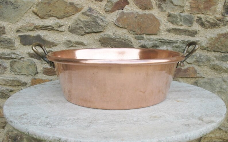 May include: A large copper bowl with two brass handles. The bowl is round and has a smooth, shiny surface. It is sitting on a light gray stone surface.