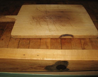 May include: A wooden cutting board with a handle. The board is worn and has many knife marks. It is sitting on a wooden surface.