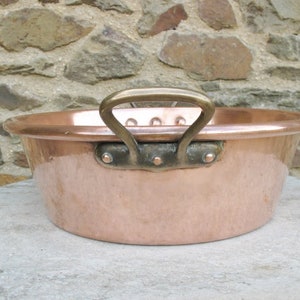 May include: A large copper pot with a single brass handle. The pot has a rounded bottom and a wide, shallow opening. The handle is attached to the pot with rivets.
