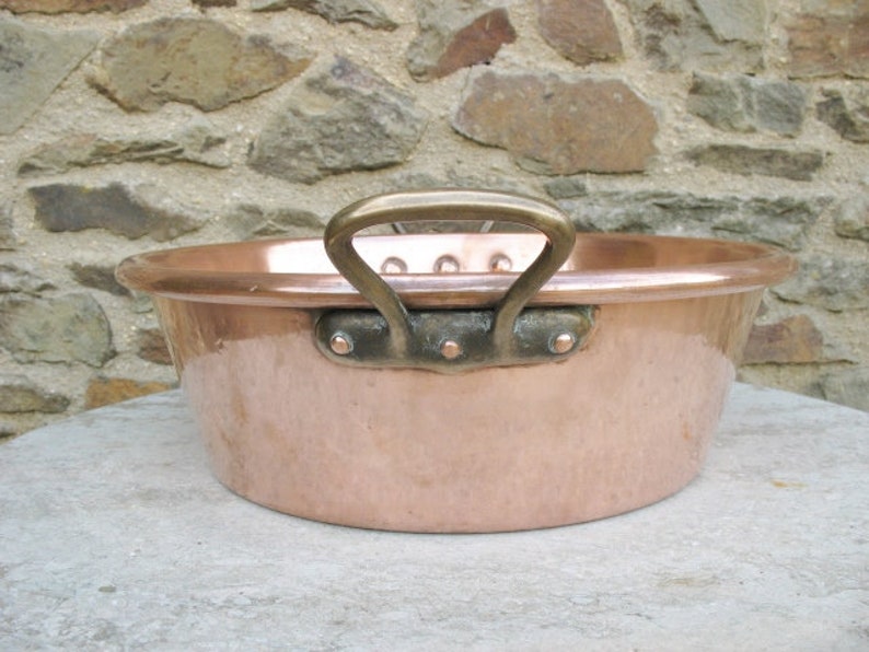 May include: A large copper pot with a single brass handle. The pot has a rounded bottom and a wide, shallow opening. The handle is attached to the pot with rivets.