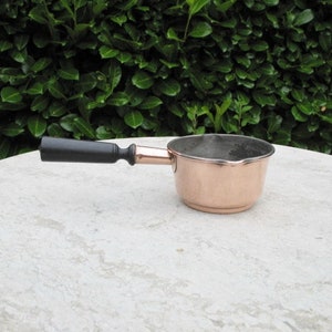 Peut inclure: Une casserole en cuivre avec une poignée en bois noir. La casserole est vide et repose sur une surface blanche.