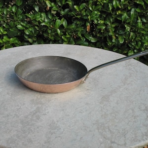 May include: A copper frying pan with a black handle. The pan is sitting on a light gray surface.