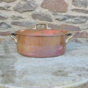 May include: An oval copper pot with brass handles and a lid. The pot is sitting on a light grey surface.