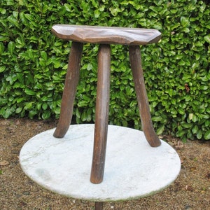 May include: A rustic wooden stool with three legs and a rounded top. The stool is made of dark brown wood and has a natural finish. The stool is sitting on a white marble table.
