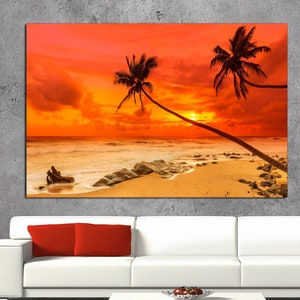 May include: A vibrant orange and yellow sunset over a tropical beach with two palm trees silhouetted against the sky. The beach is sandy with rocks and waves crashing on the shore.