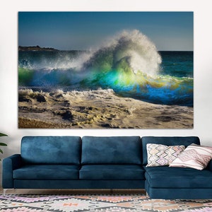 May include: A large wave crashing on a sandy beach. The wave is a vibrant blue and green color, with white foam at the crest. The beach is a light brown color, with some rocks and seaweed visible.