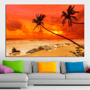 May include: A sunset over a beach with two palm trees silhouetted against the orange sky. The waves are crashing on the shore and there are rocks in the sand.
