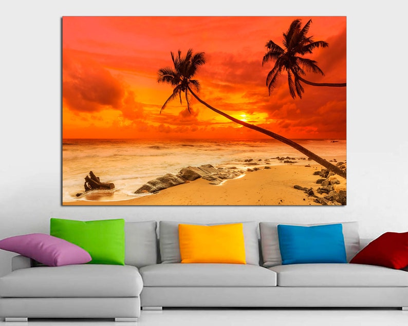 May include: A sunset over a beach with two palm trees silhouetted against the orange sky. The waves are crashing on the shore and there are rocks in the sand.