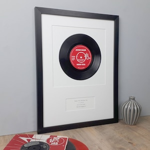 May include: Framed black vinyl record with a red label, displayed in a white mat and black frame. The label reads "Artist Name" and "Song Title". A personalized message is printed below the record.