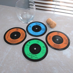 May include: Four vinyl record coasters with Adam and the Ants song titles, in orange, blue, and green. A clear glass and a cork are also present. The coasters are round and feature the labels of 45 RPM records.