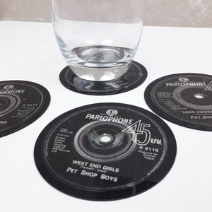 Puede incluir: Posavasos de vinilo negro con texto blanco, incluyendo "PARLOPHONE" y "WEST END GIRLS PET SHOP BOYS". Un vaso transparente descansa sobre uno de los posavasos. Los posavasos son redondos y tienen un diseño vintage.