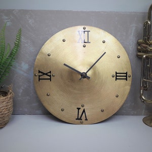 May include: A brass-colored clock made from a cymbal. The clock features black Roman numerals and black clock hands. The clock is round and has a textured surface. The clock is a unique decorative item.