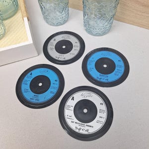 May include: Four vinyl record coasters with black edges and white or blue centres. The coasters feature song titles like "Torch" and "Say Hello, Wave Goodbye" by Soft Cell. The coasters are designed to protect surfaces from drink condensation.