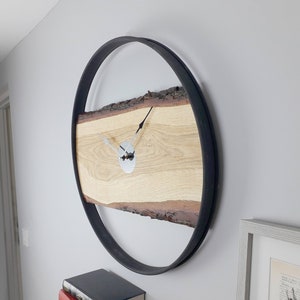 Peut inclure: Une horloge murale ronde avec un cadran en bois naturel et un cadre en métal noir. Le cadran est en bois clair avec un bord brut. Les aiguilles de l'horloge sont argentées.