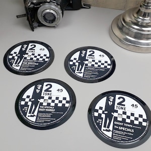 Puede incluir: Cuatro posavasos de vinilo en blanco y negro con el logotipo de la banda The Specials y títulos de canciones. Los posavasos tienen un patrón de tablero de ajedrez y el logotipo de 2 Tone Records. Una cámara vintage y una base de lámpara en el fondo.