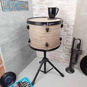 Op de afbeelding: Een bijzettafel in de vorm van een trommel met een lichte houtnerf afwerking, zwarte randen en een zwart statief. Bovenop staat een zwarte mok. Het bord erboven luidt "MUSIC ROOM". Een trompet en vinylplaten zijn ook zichtbaar.