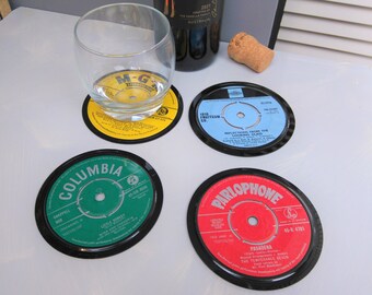 Puede incluir: Cuatro posavasos de discos de vinilo con bordes negros. Uno tiene una etiqueta amarilla con el texto "M-G", otro una etiqueta azul con "Reflections From The Looking Glass", una verde con "Columbia" y una roja con "Pasadena". También hay un vaso y una botella de vino.
