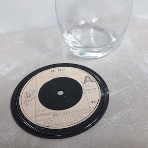 Puede incluir: Un posavasos de vinilo negro con el título de la canción "Night Fever" de los Bee Gees. El posavasos tiene un borde negro y una etiqueta blanca con texto. Un vaso transparente está encima.