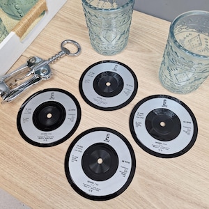 以下が含まれることがあります： 「DESIRE」と「U2」の文字が入った、黒とシルバーのビニールレコードコースター4枚。模様入りの青いグラス2つと、銀色のコルク抜きも明るい木製の表面に見えます。