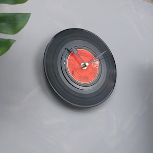 May include: A wall clock made from a black vinyl record. The clock face is a red record label with the text "HEAVEN" and "BRYAN ADAMS". The clock hands are black and the clock is mounted on a gray wall.