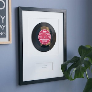 May include: A black framed wall display featuring a centered 7-inch vinyl record with a red Brunswick label. The record is mounted on a white mat board with a small rectangular cutout below containing the printed text "Forever our song, Love you, Elle x".