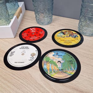 Puede incluir: Cuatro posavasos redondos hechos de discos de vinilo vintage. Cada posavasos presenta un diseño de etiqueta de disco diferente con texto y gráficos. Los posavasos son negros con etiquetas coloridas, incluyendo rojo, amarillo y blanco.