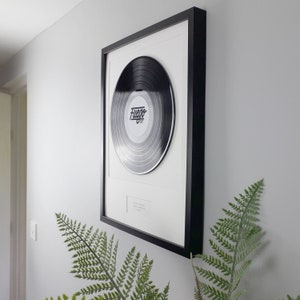 Puede incluir: Un disco de vinilo negro montado en un marco rectangular negro con un paspartú blanco. El disco tiene una etiqueta central blanca con la palabra Fuego impresa en letra cursiva negra. Una pequeña placa blanca se sitúa bajo el disco.
