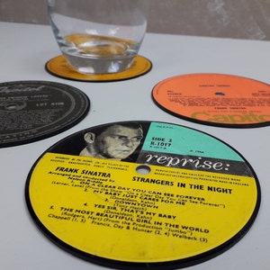 Puede incluir: Un conjunto de posavasos de discos de vinilo vintage con arte de álbumes clásicos y títulos de canciones. Un posavasos está debajo de un vaso. Los posavasos son negros, amarillos y naranjas, con texto que incluye "Frank Sinatra" y "Strangers in the Night".